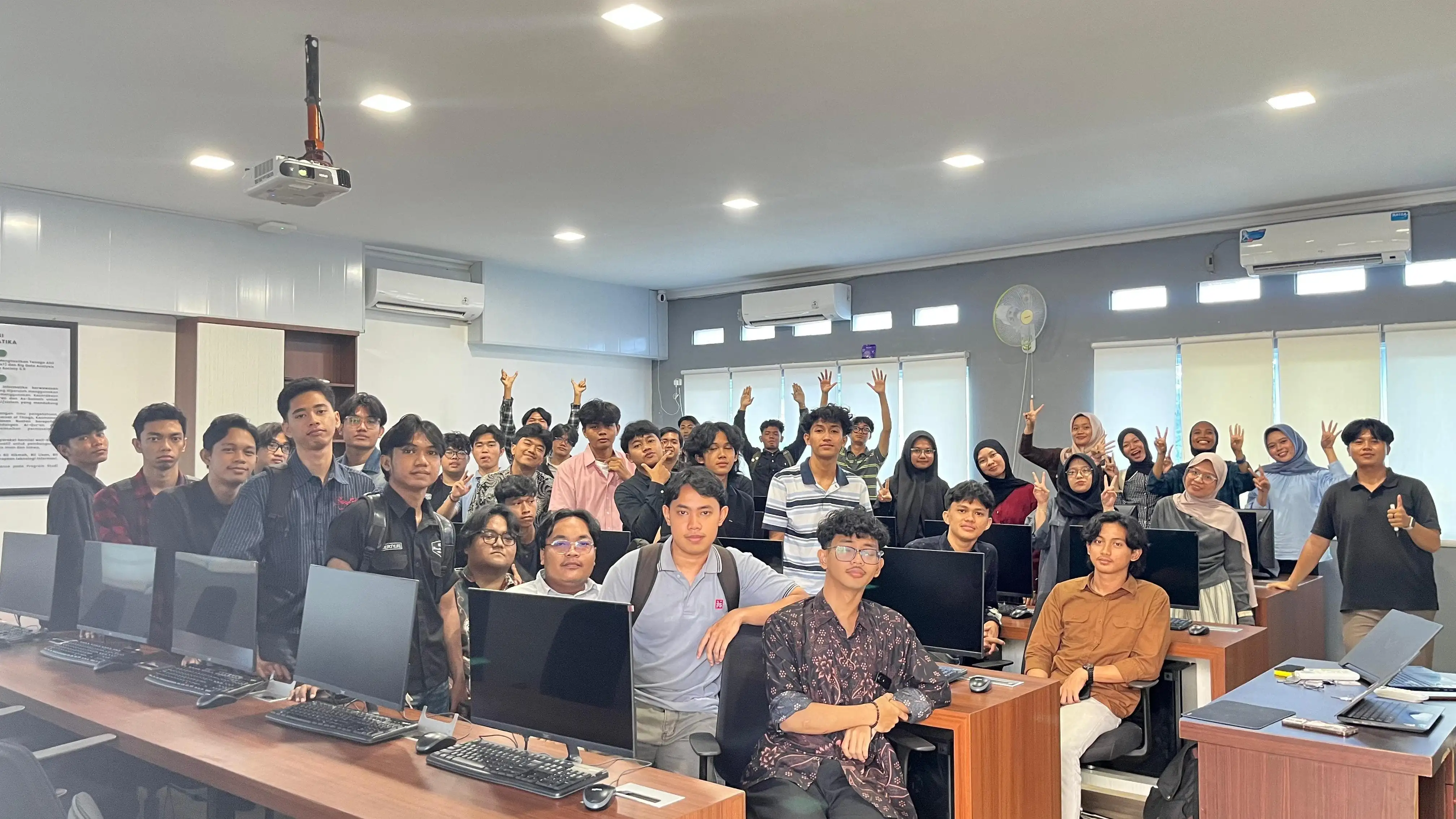 A computer lab at the end of a practicum session. Around
            thirty-five undergraduates stand and sit between two rows of
            monitors, facing the camera.